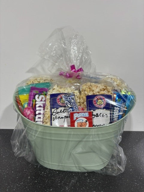 A green gift basket filled with various candies and popcorn, covered with a clear plastic wrap and tied with a pink ribbon.