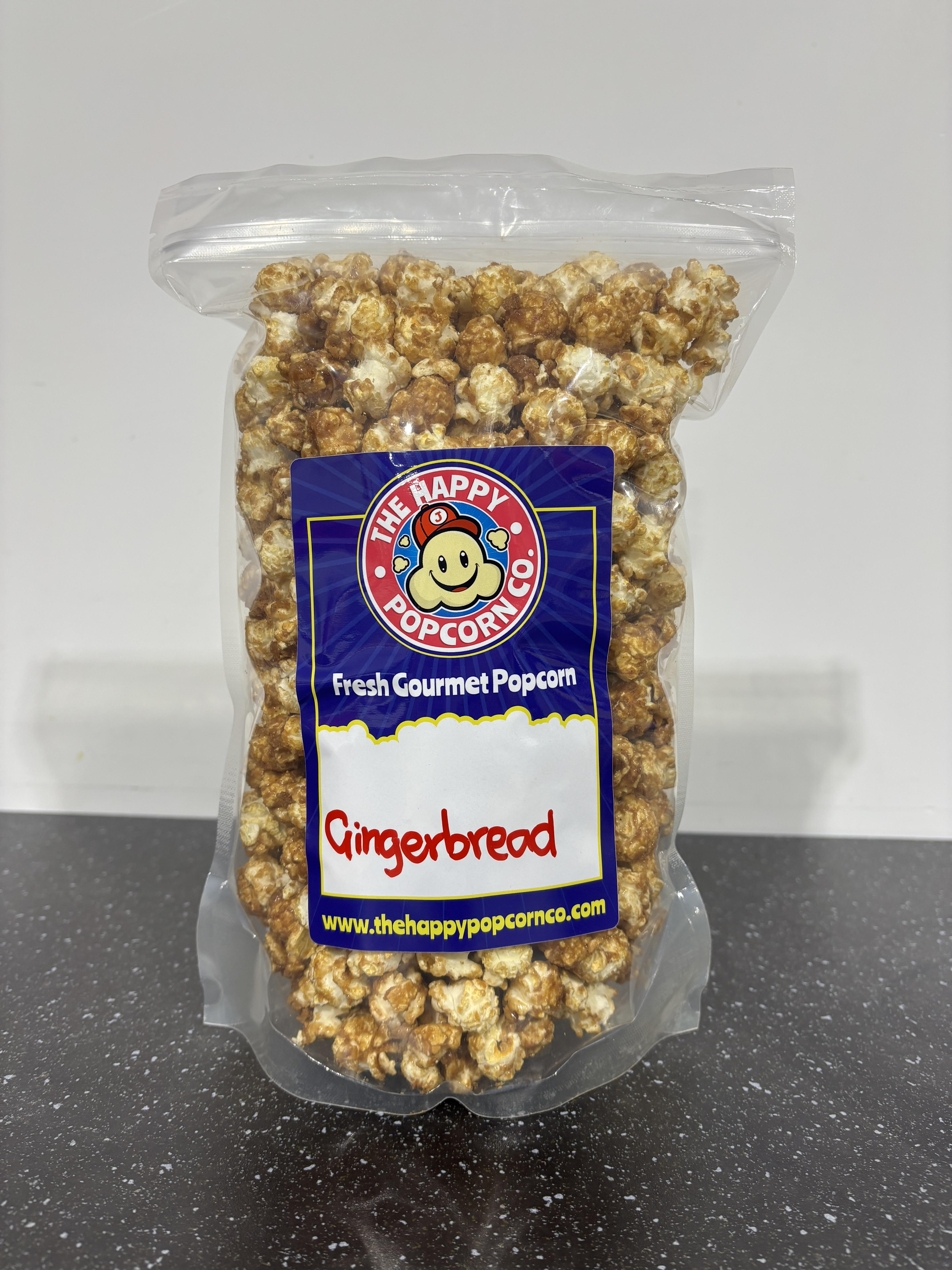 A clear plastic bag of The Happy Popcorn Co. Gingerbread flavored gourmet popcorn, showing the golden-brown coated popcorn kernels inside, set on a dark countertop against a light gray background.